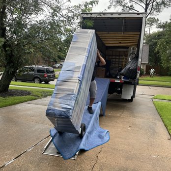 Expert Movers - A person is in the process of loading a wrapped mattress onto a dolly, positioning it to move into a large moving truck parked on a concrete driveway. The truck's back door is open, revealing moving supplies and boxes inside. The scene is set in a suburban neighborhood with trees, grass, and parked cars visible in the background. - Movers Near Me