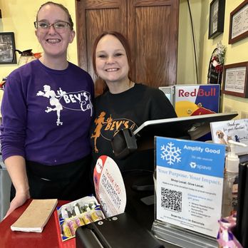 Bev's Cafe - Phee Phee and Katie are the best serving duo bar none!! Food, service, atmosphere. - Two women, one with glasses and the other with red hair, stand behind a cafe counter, smiling at the camera. They are wearing Bev's Cafe shirts, one purple and one black. The background includes a wooden cabinet with holiday decorations, framed certificates on the wall, and a sign for Red Bull. The counter has a cash register, a thank you sign, and various small items. The setting appears to be inside Bev's Cafe, a community-focused eatery. - American Near Me - Red Wing, Minnesota