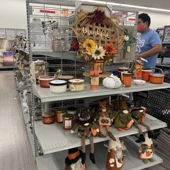 Burlington - Halloween decors - The image shows store shelves with various fall-themed decorations, including candles in jars, plush figurines of gnomes, and a floral wreath with a "thankful" sign. A person wearing a blue shirt and red face mask is visible in the background, browsing the items. The display is organized with seasonal decor items suitable for home decoration and gift-giving. - Department Stores Near Me - Lancaster, California