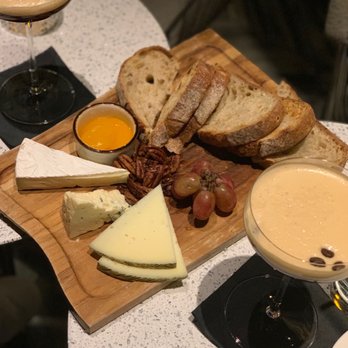 Le Bar - Charcuterie - A cheese and bread platter features various cheeses, grapes, nuts, honey, and sliced bread on a wooden board. Two cocktails and a glass of wine are placed nearby on a speckled table, with black napkins underneath the drinks. The setting appears to be a restaurant or bar with a casual dining atmosphere. - Bars Near Me - Chicago, Illinois