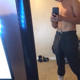 A young man is shirtless, smiling, and taking a mirror selfie in a bedroom. He wears a backwards cap, dark sweatpants, and socks, with shoes on the floor behind him.