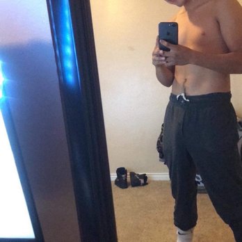 Mon Chalet - A young man is shirtless, smiling, and taking a mirror selfie in a bedroom. He wears a backwards cap, dark sweatpants, and socks, with shoes on the floor behind him. - Adult Entertainment Near Me - Aurora, Colorado