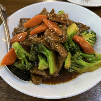 Veggie House - A white ceramic bowl holds a stir-fried dish with broccoli florets, sliced carrots, and pieces of beef coated in a dark savory sauce. The dish is served on a wooden table, with a metal spoon resting on the side of the bowl. The vegetables are cooked but retain their color and texture, and the sauce appears glossy and flavorful. - Vegan Near Me - Chicago, Illinois