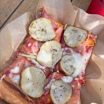 Rosetta Bakery - A close-up of a pizza slice with potato slices, ham, melted cheese, and sprinkled herbs, served on parchment paper. - Bakeries Near Me - Miami, Florida