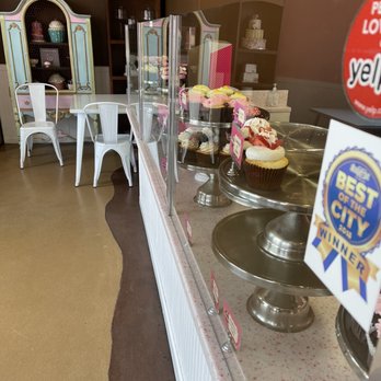 Smallcakes A Cupcakery - Cupcakes are arranged on metal stands behind a glass display case in a bakery or cafe environment. - Bakeries Near Me - Albuquerque, New Mexico