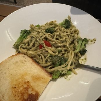 Harrison’s - A white round plate contains a serving of pasta mixed with broccoli florets, cherry tomato pieces, and pesto sauce. A silver spoon rests on the plate's edge. A slice of toasted bread is placed on the plate's side. The background shows part of a person's arm and a table surface, indicating a dining setting. - Steakhouses Near Me - Indianapolis, Indiana