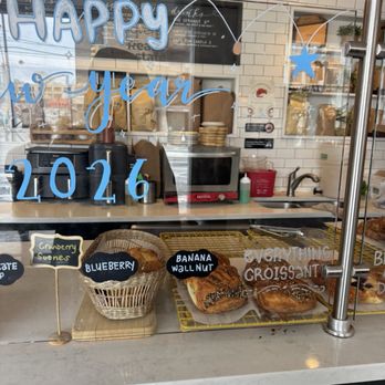 Concept Coffee - A bakery display case features various pastries such as blueberry scones, banana walnut bread, and croissants. The pastries are arranged on cooling racks and a wooden board. The glass window has handwritten labels and New Year's decorations, including a sign that says 'HAPPY new year 2026' with stars and balloons. The background shows a bakery interior with kitchen equipment and white tiled walls. - Coffee & Tea Near Me - Queens, New York