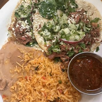 Tune-Up Cafe - Lamb tacos - A white plate holds lamb tacos garnished with chopped onions, cilantro, and leafy greens, accompanied by a serving of seasoned rice, refried beans, and a small cup of salsa. The setting suggests a casual dining environment at Tune-Up Cafe in Santa Fe, NM, known for fusion cuisine including Salvadoran and New Mexican dishes. - New Mexican Cuisine Near Me - Santa Fe, New Mexico