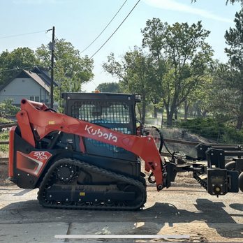 MK Equipment - Getting yard ready for sod - A Kubota SVL75-3 compact track loader with a front attachment is parked on a concrete surface outdoors. The loader has a black cab with a protective grid, rubber tracks, and a red arm with the Kubota logo. The front attachment appears to be a brush cutter or similar implement. The background features trees, a house, and utility poles, indicating a rural or semi-rural setting. - Excavation Services Near Me - Fruita, Colorado