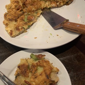 Somac - Seafood Pancake - The image features two white plates with Korean pancakes. The larger plate holds a thick, irregularly shaped pancake with visible green onions and other ingredients, accompanied by a metal spatula. The smaller plate contains a piece of pancake with a crispy, golden-brown exterior, showing green vegetables and possibly meat inside. The pancakes are served on a dark wooden table. - Chicken Wings Near Me - Chantilly, Virginia