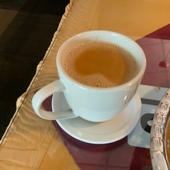 AAP India - 5. Special Tea - A white ceramic cup filled with tea sits on a matching saucer on a wooden table, with a basket partially visible nearby. - Indian Near Me - Cincinnati, Ohio