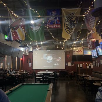 Third Base - Huge screen games and a pool table. - The image shows the interior of a sports bar with a pool table in the foreground, surrounded by tables and chairs. Hanging from the ceiling are multiple sports flags and string lights. A large screen at the back displays a commercial for AT&T. The bar has a dark wood floor and walls decorated with sports memorabilia and framed pictures. There are a few patrons seated at tables, and the overall atmosphere appears casual and entertainment-focused. - Dive Bars Near Me - Chandler, Arizona