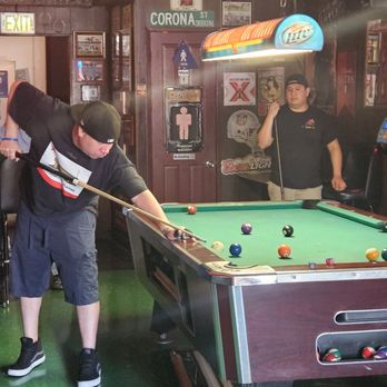 R Place - A young man is taking a shot at a pool table in a bar decorated with vintage posters and signs, with another person standing nearby. - Dive Bars Near Me - Pasadena, California