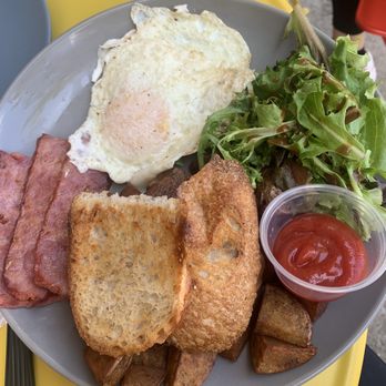 Belly Cake - Farmer's Plate - A breakfast plate includes a fried egg with a runny yolk, several strips of bacon, two slices of toasted bread, and crispy hash browns. A side salad with mixed greens and a small cup of ketchup is also present. The meal is served on a gray plate placed on a yellow table, with part of a gray plate visible in the background. - Pancakes Near Me - Bayside, New York