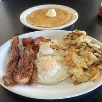 Pete's Cafe - A breakfast plate featuring bacon, fried eggs, and hash browns, with a side of pancakes topped with butter in the background. - Breakfast & Brunch Near Me - Thornton, Colorado