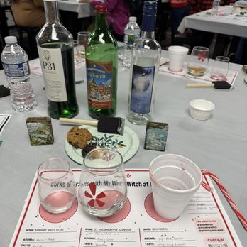 Whimsy & Wine - A table at a wine tasting event features three bottles of wine, several glasses, snacks, and tasting notes on a placemat. The background shows multiple people seated at tables, engaged in tasting and socializing. The table includes water bottles, a small plate with snacks, and tasting sheets with handwritten notes about the wines. - Home Decor Near Me - Fraser, Michigan