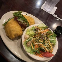 A plate holds a sandwich with lettuce, tomato, and meat, alongside a mixed salad with vegetables and shredded cheese, with a glass of water nearby.