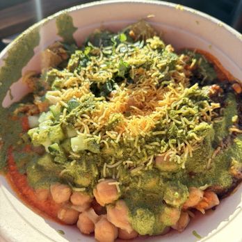 RASA - A close-up of a bowl containing chickpeas, topped with chopped vegetables, shredded cheese, and a green sauce or chutney. The dish appears to be served in a disposable bowl, with sunlight illuminating the food. The ingredients suggest a flavorful, seasoned dish, possibly from Indian or Middle Eastern cuisine. - Indian Near Me - Fairfax, Virginia