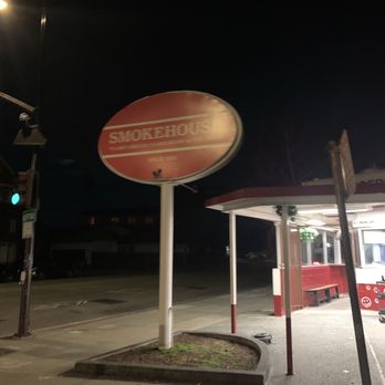 Smokehouse - A red and white oval Smokehouse sign is mounted on a white pole outside a building at night. The sign is illuminated but the text is not clearly visible. Nearby, there is a small shelter with a bench and a street corner with traffic lights. The area appears to be a quiet street at night with minimal lighting. - Fast Food Near Me - Berkeley, California