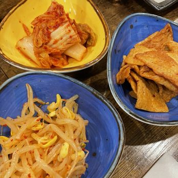 Jang Dok Dae - Three bowls of Korean banchan, including kimchi, bean sprouts, and fried tofu, are arranged on a wooden table. - Korean Near Me - Flushing, New York