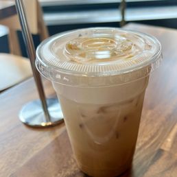 Dolce De Leche Iced Latte - A plastic cup filled with iced latte, featuring a clear lid and a straw, placed on a wooden table in a cafe setting.