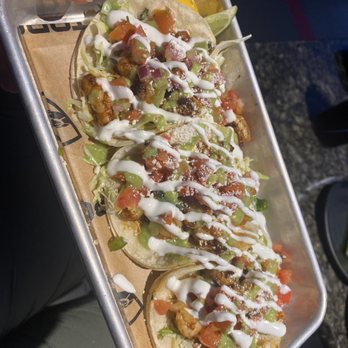 Topgolf - Shrimp tacos!!! - A rectangular metal tray contains several shrimp tacos with soft tortillas filled with cooked shrimp, diced tomatoes, lettuce, and sauce. The tacos are topped with a drizzle of sour cream and sprinkled with cheese or seasoning. A lemon wedge is visible at the edge of the tray, and the tray is lined with paper. The background suggests an outdoor setting, possibly at a food event or restaurant. - Golf Near Me - El Paso, Texas