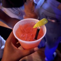 tequila sunrise - A hand holds a pink-colored cocktail with a lemon wedge and straw, likely in a bar or social setting with people in the background.