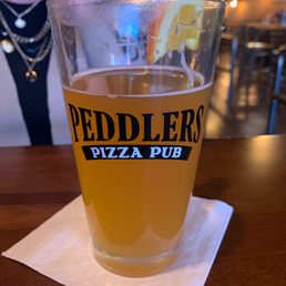 Blue Moon - A pint glass filled with beer, garnished with a lemon wedge, bearing the Peddlers Pizza Pub logo, placed on a napkin on a wooden table.