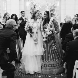 The image depicts a wedding ceremony with two women at the center, one in a white wedding dress holding a bouquet, and the other in traditional attire with floral garlands. They are smiling at each other, surrounded by seated and standing guests, some clapping and smiling. The setting appears to be indoors with large windows and floral decorations in the background.