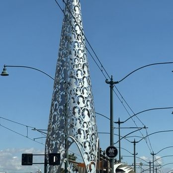 Downtown Mesa - Sculpture - A tall, metallic sculpture with a perforated, abstract design is situated in an urban setting, surrounded by streetlights and tram wires. - Art Museums Near Me - Mesa, Arizona
