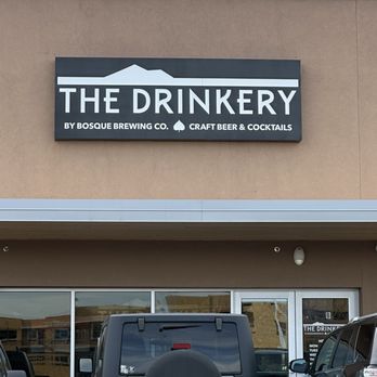The Drinkery - Entry sign - A rectangular sign for The Drinkery is mounted on the exterior wall of a beige building. The sign features the business name in large white letters on a dark background, with a stylized roof graphic above. Below, smaller text indicates it is operated by Bosque Brewing Co. and offers craft beer and cocktails. The parking lot in front contains several vehicles, including a white Jeep with a spare tire cover, and the entrance has glass doors and windows. - Beer Bar Near Me - Santa Fe, New Mexico