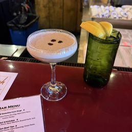 Espresso Martini - A cocktail served in a coupe glass is topped with a frothy layer and garnished with three coffee beans. Next to it, a tall green glass contains ice and lemon slices. The setting appears to be a bar with a menu partially visible and a background featuring bar supplies and a wooden cabinet.
