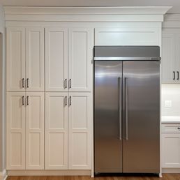 Pantry - The image shows a kitchen with white, paneled cabinetry on the left and right sides, with black handles. A large stainless steel refrigerator with vertical handles is positioned centrally, integrated into the cabinetry. The background includes a white wall with a power outlet and a section of countertop visible on the right side. The flooring appears to be wood.