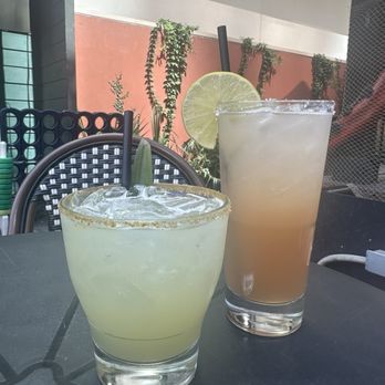 Augustine Cocktail Lounge - Two cocktails with lime slices are placed on a dark outdoor table at Augustine Cocktail Lounge, featuring indoor and outdoor seating with plants in the background. - Cocktail Bars Near Me - Austin, Texas
