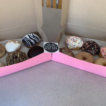 North Fork Doughnut - Jelly filled, apple fritter, apple crumb, caramel crumb cake amongst others - A pink and gray box contains a variety of donuts with different toppings, including sprinkles, icing, and glaze, on a gray table. - Donuts Near Me - Mattituck, New York