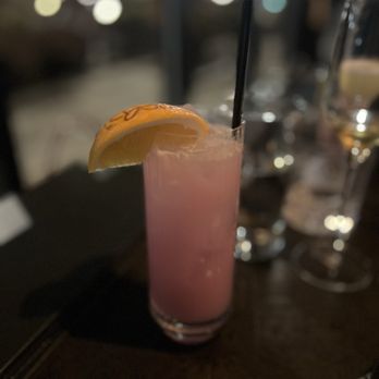 Dearly Beloved - Soda Pop - A pink-colored cocktail in a tall glass with a lemon wedge garnish, set on a dark table in a dimly lit bar or restaurant. - Cocktail Bars Near Me - Chicago, Illinois