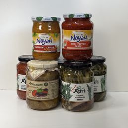 Noyan - Six glass jars of preserved foods are arranged on a white surface against a plain white background. The jars contain eggplant caviar, red pepper paste, marinated chervil, and grilled vegetables. Each jar has a label with branding and product information, with some jars featuring decorative cloth covers secured with string. The image showcases a variety of preserved Armenian-style vegetables and spreads.