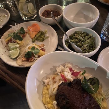 Penca - The image shows a table set with Mexican dishes, including tacos garnished with lime, cilantro, and vegetables, a bowl of chopped green vegetables, and a large meat portion served over a bed of yellow rice or polenta. There are also condiments, a glass of water, and various glasses and bowls on the table, indicating a meal setting in a restaurant. The setting suggests a casual dining environment with a focus on traditional Mexican cuisine. - Mexican Near Me - Tucson, Arizona