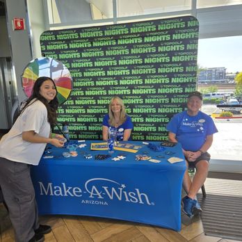 Topgolf - A young woman stands at a Make-A-Wish Arizona event table, smiling at the camera. The table is covered with a blue tablecloth featuring the Make-A-Wish Arizona logo and has various promotional items, including pens, wristbands, and papers. Two volunteers, a woman and a man, are seated behind the table, both wearing blue shirts with the Make-A-Wish logo. The background includes a large window with a view of a parking lot and buildings outside, and a backdrop with the words 'Nights' and 'Topgolf' repeated in green and black. A colorful spinning wheel is positioned behind the table. - Golf Near Me - Gilbert, Arizona