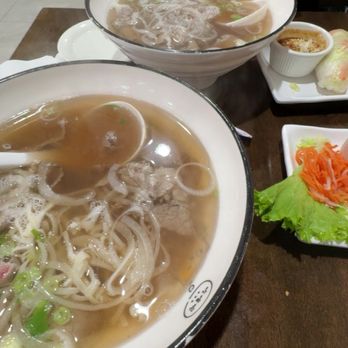 Pho Table - Bath Beach - Two bowls of Vietnamese pho with clear broth, rice noodles, and green onions are placed on a dark wooden table. One bowl is closer to the camera, and the other is further back, with steam rising from both. Side dishes include a small bowl of dipping sauce, a plate with lettuce and shredded carrots, and a rice paper roll. A person in a black shirt is visible in the background, holding a smartphone. The setting appears to be a casual Vietnamese eatery. - Vietnamese Near Me - Brooklyn, New York