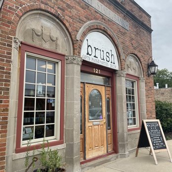 Brush Home Decor and Paint Studio - The image shows the exterior of Brush, a gift, paint, parties, and decor store, situated in a brick building. The storefront features two large windows with red trim, a wooden door with decorative glass panels, and a sign above that reads 'brush' with services listed below. A sidewalk chalkboard sign is placed outside, and a small plant cart with potted plants is nearby. The building has a sign indicating it is part of Cary State Bank. - Furniture Stores Near Me - Cary, Illinois