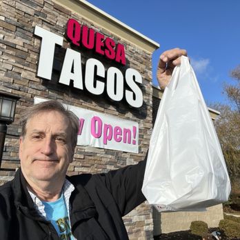 Quesa Tacos - Carry out breakfast tacos - A man is taking a selfie outside Quesa Tacos, holding a white plastic takeout bag in his right hand. The restaurant's sign, displaying the words 'Quesa Tacos' and an 'Open!' banner, is visible on a stone facade behind him. The sky is clear and blue, and there are trees with autumn foliage in the background. - Mexican Near Me - Carmel, Indiana