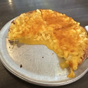Apollo Pizza - A slice of macaroni and cheese pizza with melted cheese and cooked pasta on top is placed on a round metal tray. The pizza has a golden-brown crust and cheese that is slightly browned. The tray is on a dark wooden surface, and the background is blurred. The pizza appears to be a casual dining item, featuring a combination of cheese, pasta, and pizza crust. - Pizza Near Me - Berea, Kentucky