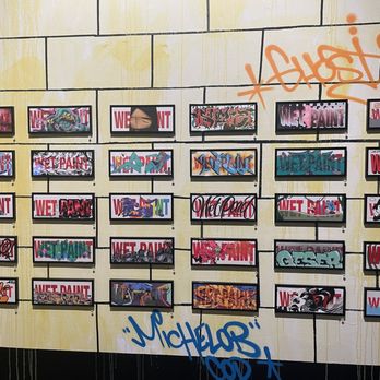 Redline - The image shows a display of small framed graffiti art pieces arranged in a grid on a yellow wall. The wall has graffiti tags and spray paint markings, including an orange tag at the top and a blue tag at the bottom. The framed artworks feature various graffiti styles, characters, and text, with some overlapping the frames. The setting appears to be an indoor art exhibit or gallery focused on street art. - Art Galleries Near Me - Denver, Colorado