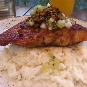 Para Maria - Salmon with guava sauce and coconut rice - A food presentation with a grilled fish fillet topped with chopped onions and crispy meat bits, placed on a bed of mashed potatoes. The plate is set on a wooden table with a glass of water and a yellow drink in the background, suggesting an outdoor dining setting. The image emphasizes the texture and garnishes of the dish. - Wine Bars Near Me - Boston, Massachusetts