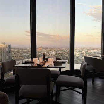 The Compass - View - A dining table with four chairs is arranged near large floor-to-ceiling windows in a high-rise restaurant. The table has cups, napkins, and a small sign that reads 'RESERVED.' The view through the windows shows a cityscape with buildings and a sky with clouds illuminated by the setting sun. The interior features dark flooring and modern furniture. - American Near Me - Phoenix, Arizona