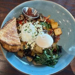 Market hash - A breakfast dish on a blue ceramic plate includes two poached eggs garnished with chopped chives, a slice of toasted bread, roasted potatoes, sautéed vegetables such as spinach and mushrooms, and a small metal cup of syrup. The plate is placed on a wooden table.