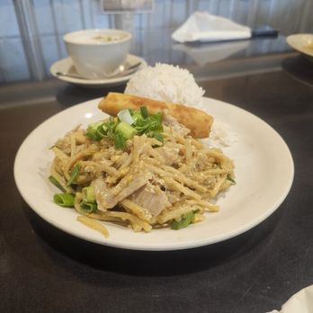Thai Kitchen - A white plate holds stir-fried noodles with pork and chopped green onions, alongside a spring roll and a mound of white rice. In the background, there is a bowl of soup on a saucer, a tissue, and a blurred restaurant setting with a metal wall and a small sign. The table surface is dark, and the overall scene suggests a casual dining environment. - Thai Near Me - Austin, Texas
