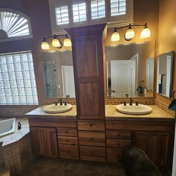 Bubbles Cleaning Service - prior to clean - The image shows a bathroom with a double sink vanity, featuring two mirrors and wall-mounted light fixtures above each mirror. The vanity has wooden cabinetry with multiple drawers and a central cabinet. A window with shutters is positioned above the vanity, allowing natural light into the space. To the left, part of a bathtub with a tiled surround is visible, along with a glass block window providing additional light. The floor is tiled, and a dog is partially visible in the lower right corner. - Home Cleaning Near Me