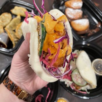 Take a Bao Eats - Crispy spicy shrimp bao - A steamed bao bun filled with fried fish, shredded purple cabbage, cucumber slices, and orange sauce, held in a person's hand. In the background, there are black plastic containers with fried dumplings and powdered pastries, along with small cups of sauce, on a dark table surface. - Asian Fusion Near Me - Chicago, Illinois
